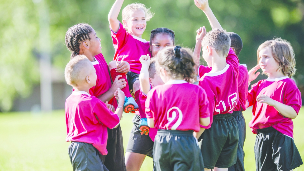 A multi-ethnic group of elementary age children are standing together in a row after their soccer game. They are cheering and holding up their teammate.